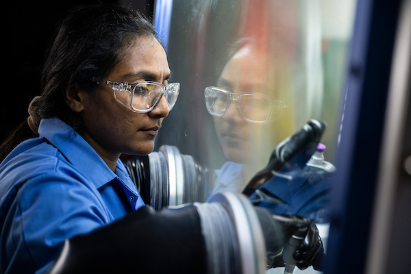 A scientist in safety goggles works intently at a lab glove box, their reflection visible on the glass